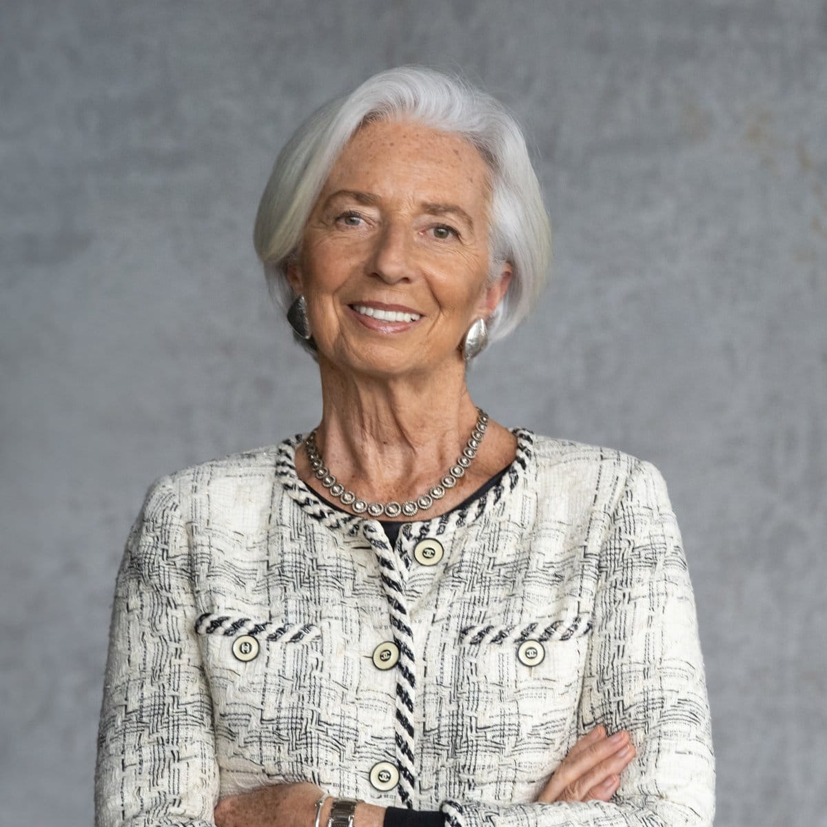 Portrait of Christine Lagarde smiling with arms crossed, wearing a white tweed jacket and silver necklace.