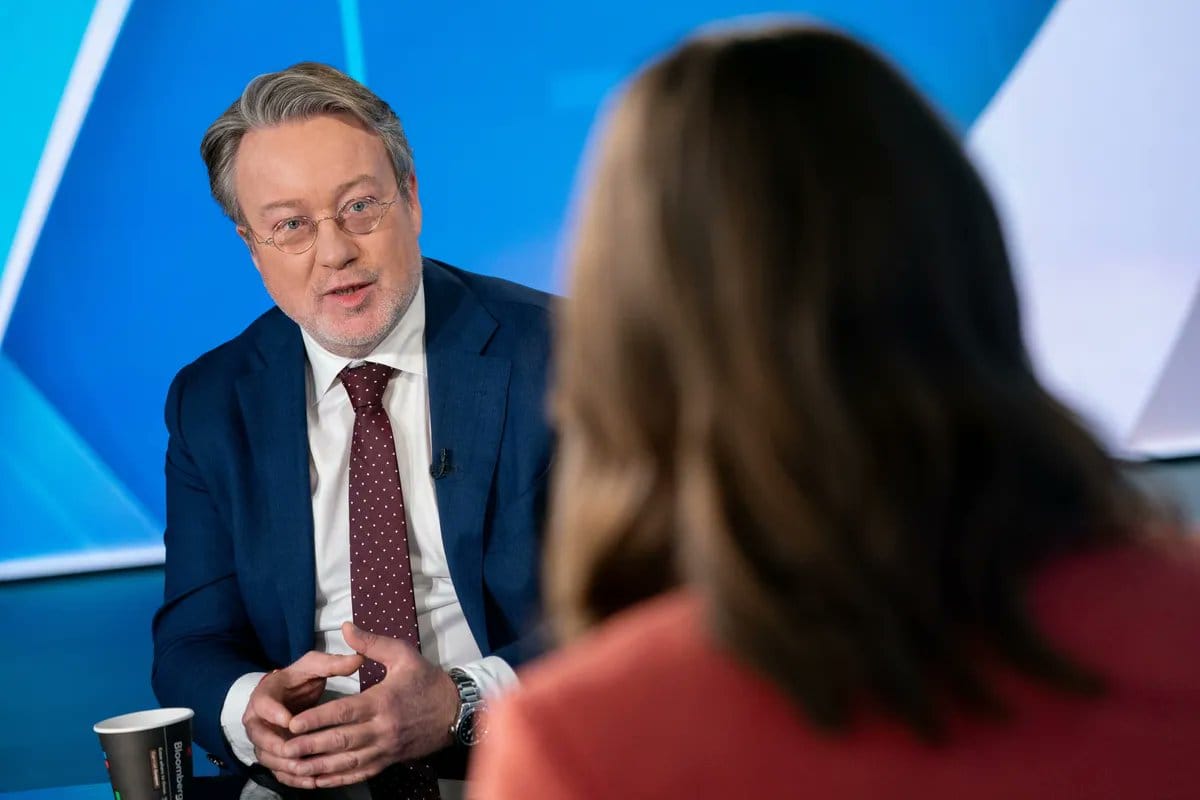 Man in blue suit with glasses and polka-dot tie speaking during interview, woman in red jacket visible from behind in studio.