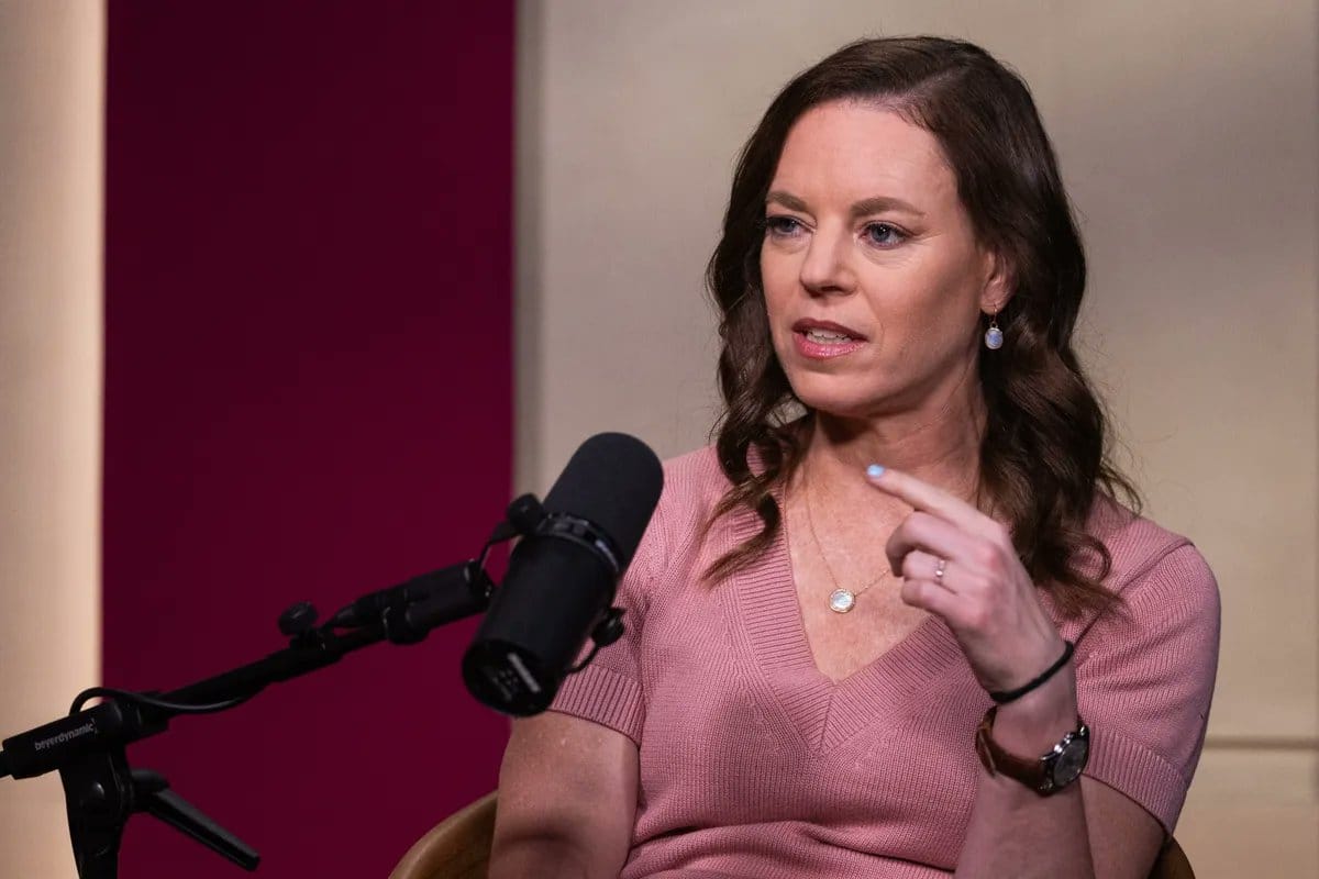 Woman with brown wavy hair in a pink sweater seated and speaking into a black microphone.