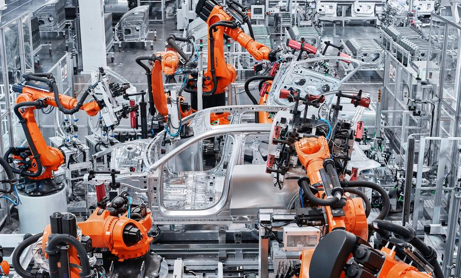 Orange robotic arms assembling a silver car chassis in a large automated factory.