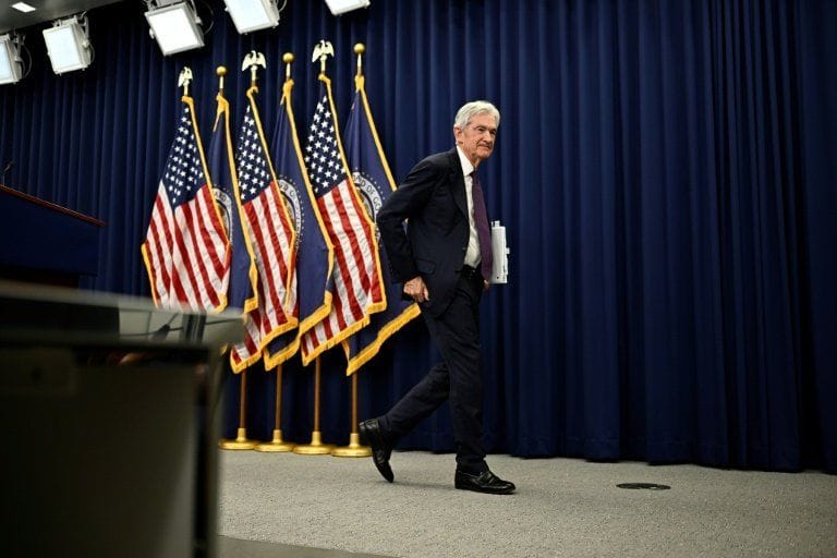 Jerome Powell in a dark suit walking in a formal room with American flags and blue curtains.