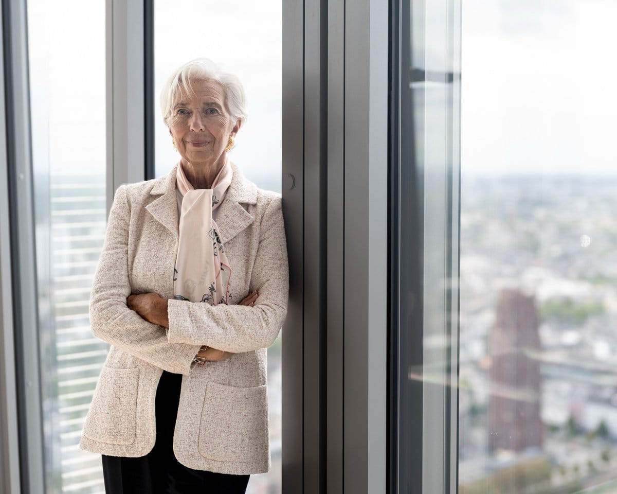 Christine Lagarde with arms crossed, standing beside a large window with a cityscape view.