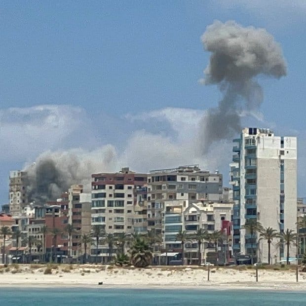 Coastal city with beach, palm trees, and multi-story buildings under a plume of dark smoke rising into blue sky.