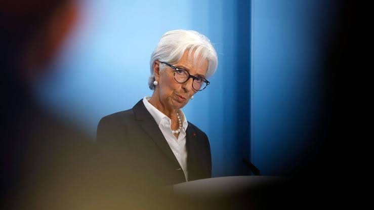 Christine Lagarde with short white hair and glasses, wearing a dark blazer over a white shirt with a pearl necklace, standing at a podium against a blue background.