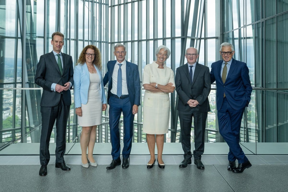 Six formally dressed individuals standing in a line in a modern room with large glass windows overlooking a city.
