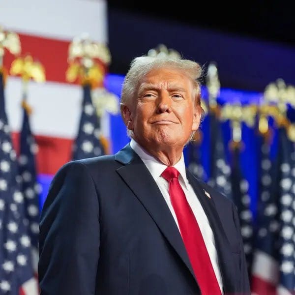 Donald Trump in a dark suit and red tie, standing before American flags.