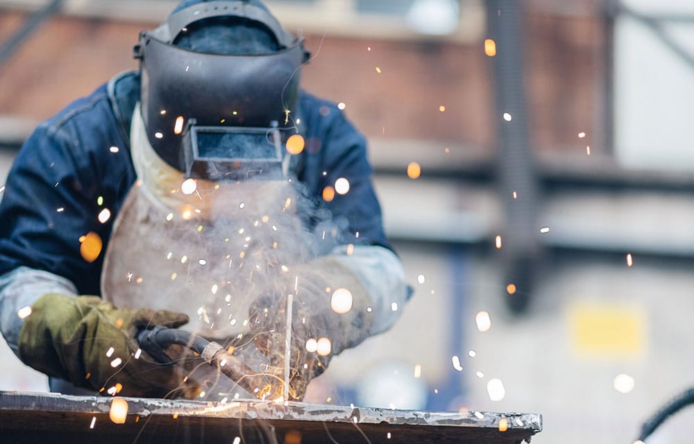 Welder in helmet, apron, and gloves operating a torch on metal with sparks flying in an industrial setting.