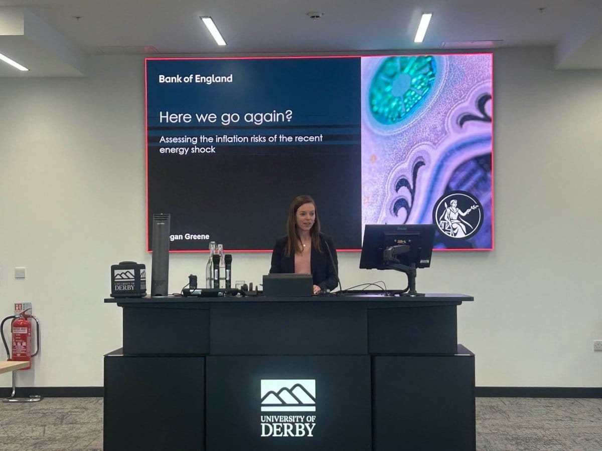 Woman at a podium with a large screen displaying a Bank of England presentation on energy shock inflation risks.