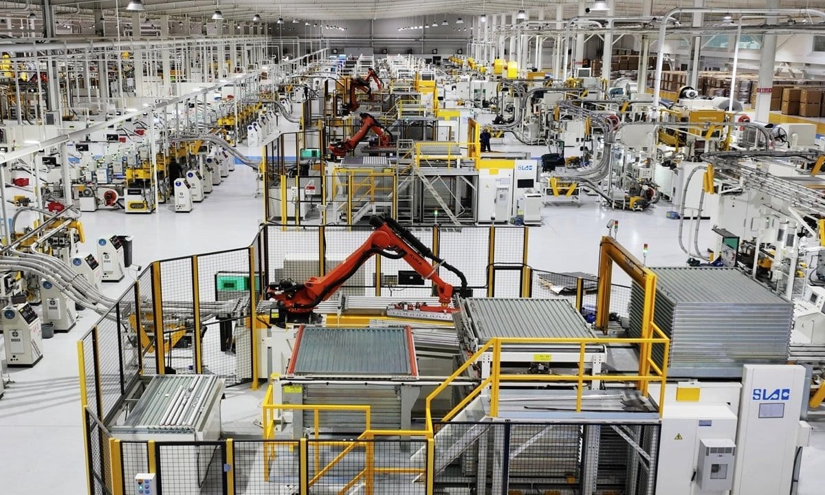 Wide interior view of a large automated factory with multiple robotic arms, conveyor systems, and production machinery.