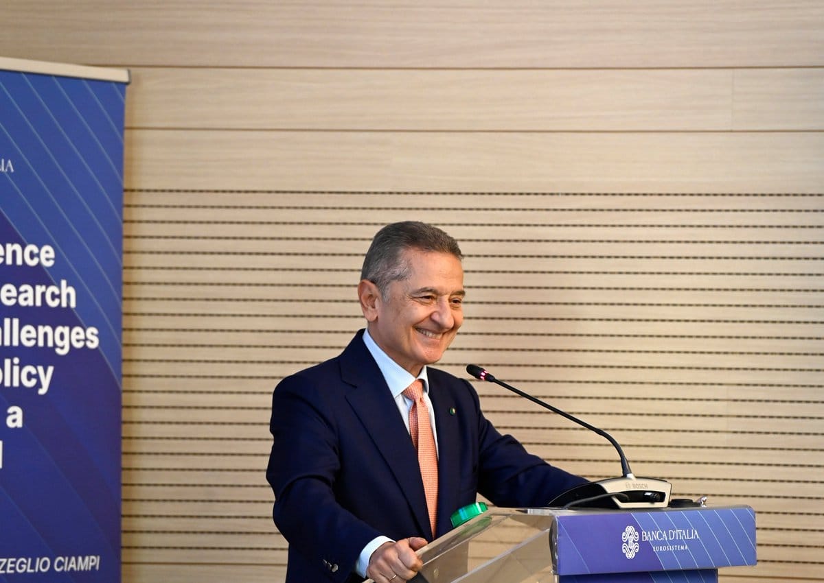 Smiling man in navy suit and orange tie at transparent podium with Banca d'Italia logo and microphone.
