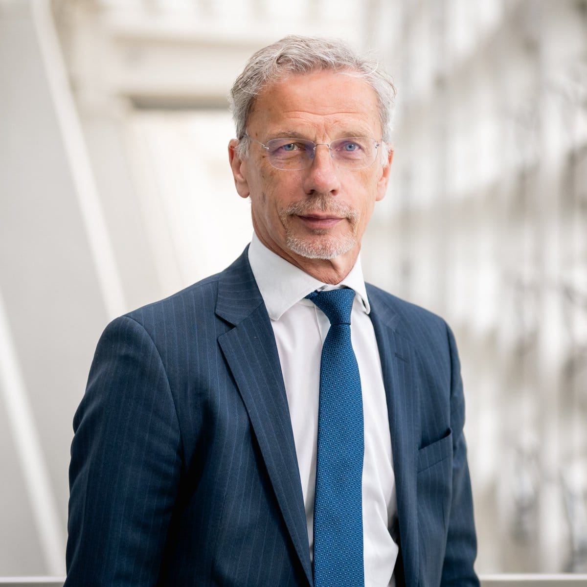 Head-and-shoulders portrait of a man with short gray hair, light beard, thin-rimmed glasses, dark blue pinstripe suit, white shirt and blue patterned tie against a blurred modern interior...