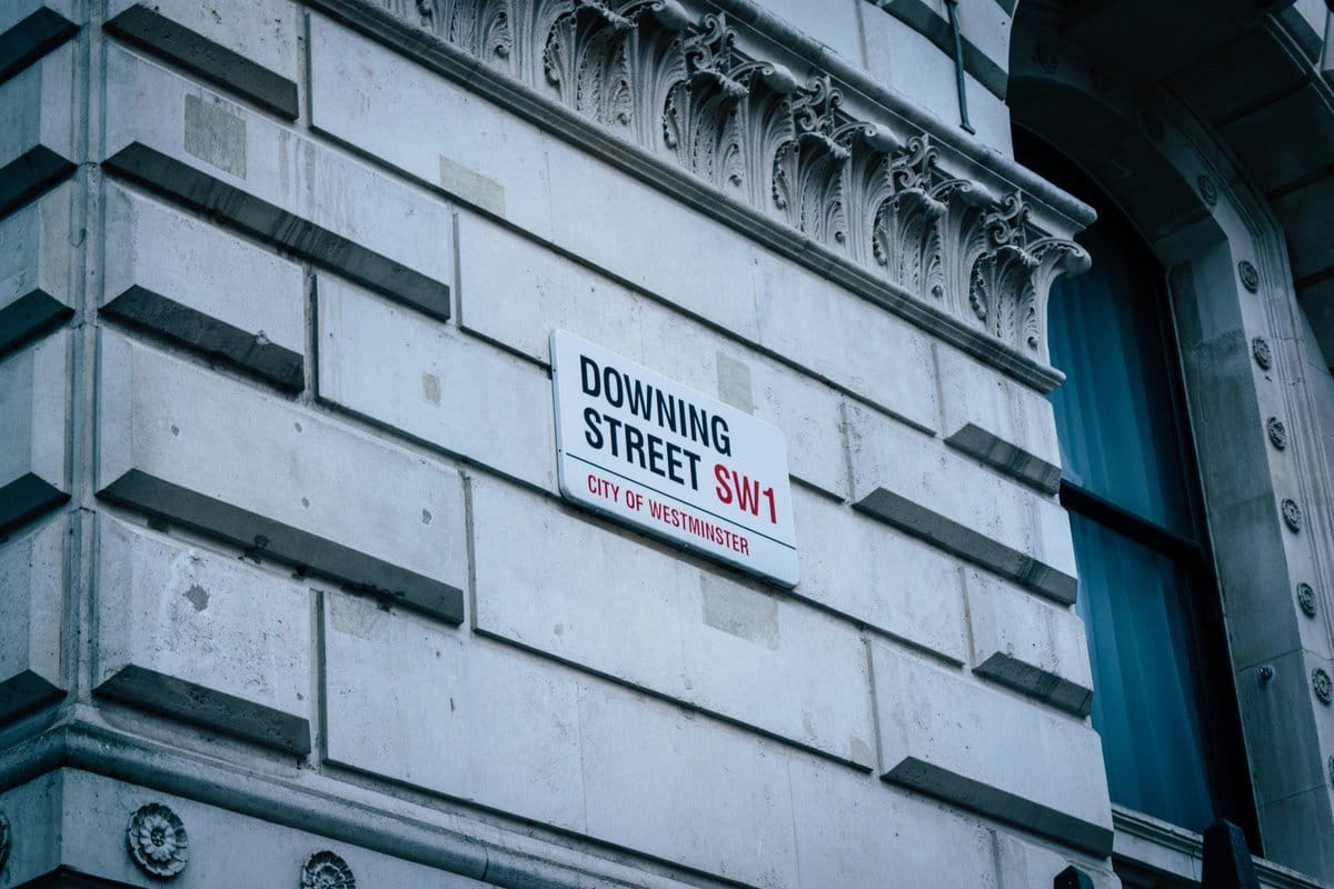 Close-up of a white sign on a stone building reading 'DOWNING STREET SW1' in black and red, with 'CITY OF WESTMINSTER' below.