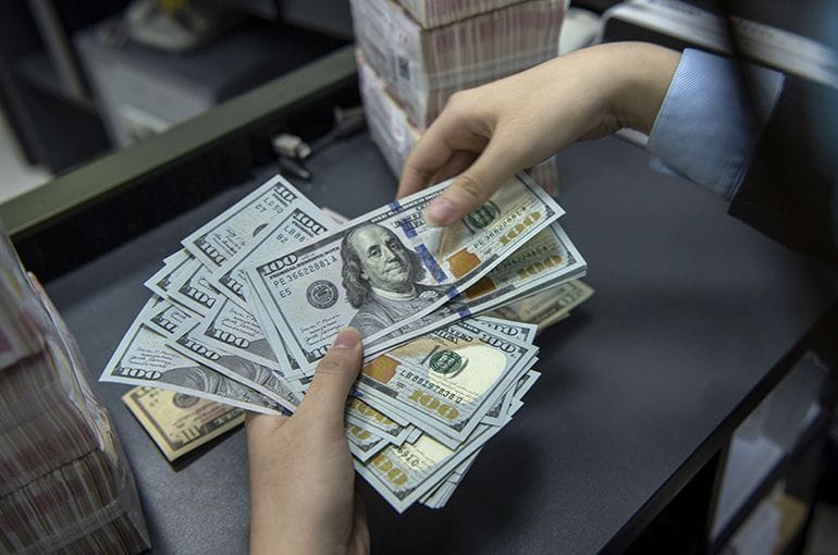 Hands counting a thick stack of US 100-dollar bills on a counter, with more bundled cash visible in the background.