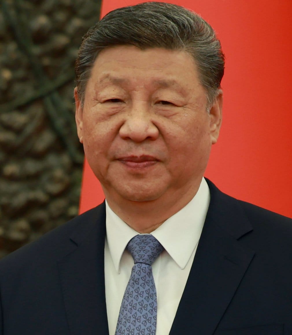 Portrait of Xi Jinping in a dark suit and patterned tie against a red Chinese flag.