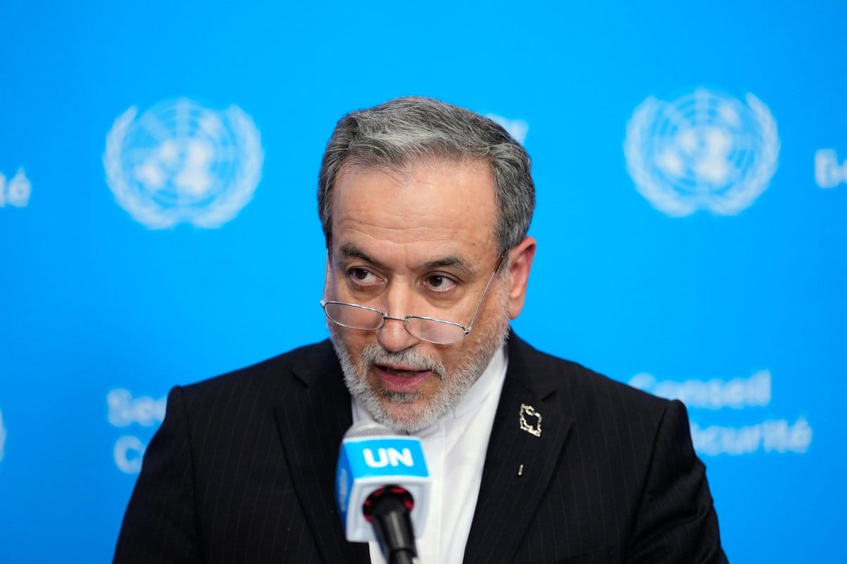 Man with gray hair, beard and glasses in a dark pinstripe suit speaking into a UN-branded microphone against a blue UN backdrop.