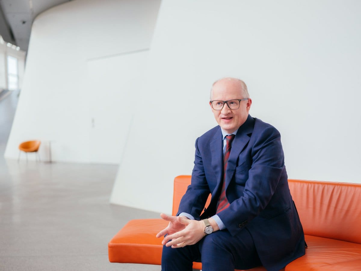 Bald man with glasses in a navy suit and striped tie sits on an orange sofa in a modern white interior, gesturing with both hands.
