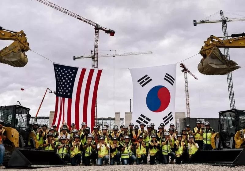 Workers in yellow vests and hard hats pose at a construction site with U.S. and South Korean flags, cranes, and excavators.