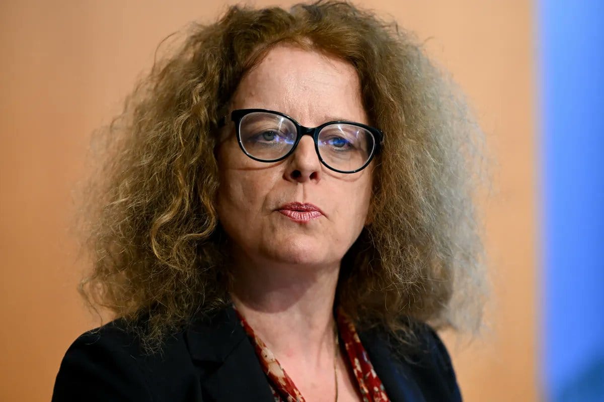 Close-up portrait of a woman with curly reddish-blonde hair and black-rimmed glasses in a dark blazer against a peach background.
