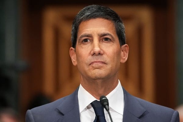 Man in a dark blue suit and navy tie speaking at a microphone, wooden doors in the background.