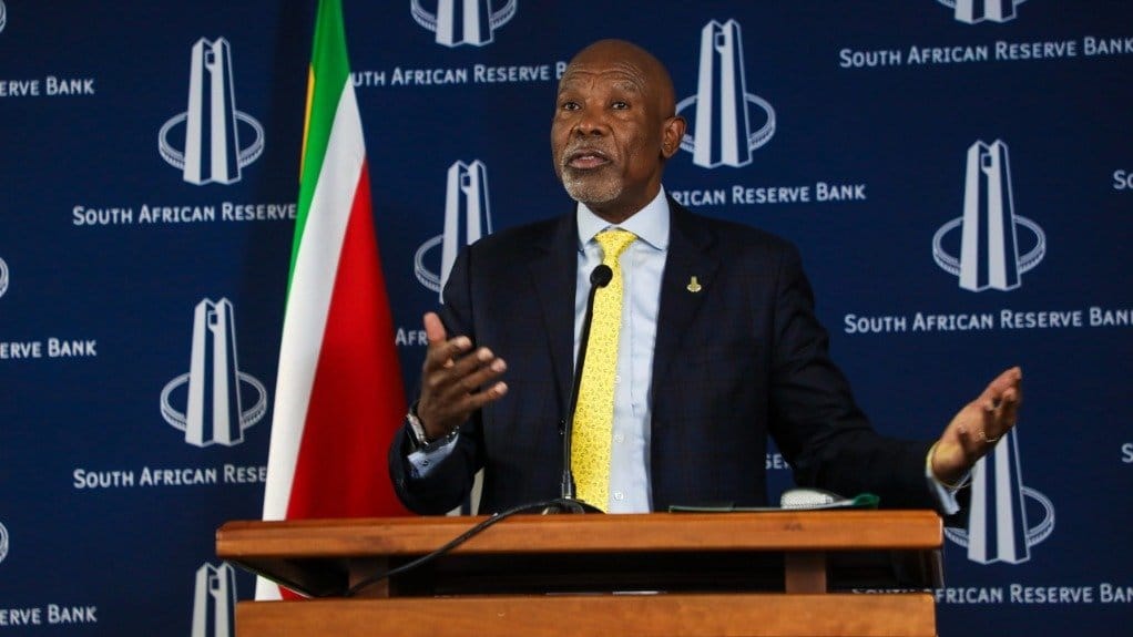 Man in a dark suit and yellow tie speaking at a wooden podium, with South African Reserve Bank logos and a South African flag behind him.