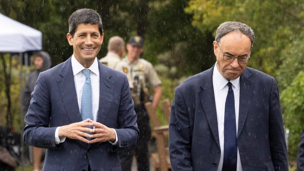 Two men in dark suits walk outdoors in rain; one smiles with hands clasped, the other looks down; a uniformed officer stands behind them.