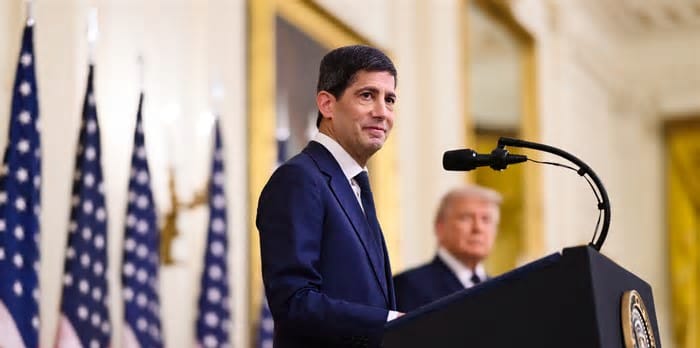 Man in a navy suit at a podium with U.S. flags; another man in a suit stands out of focus in the background.