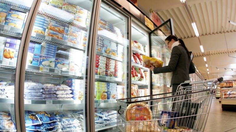 Woman at supermarket glass-door freezers selecting a frozen package, shopping cart in the aisle.