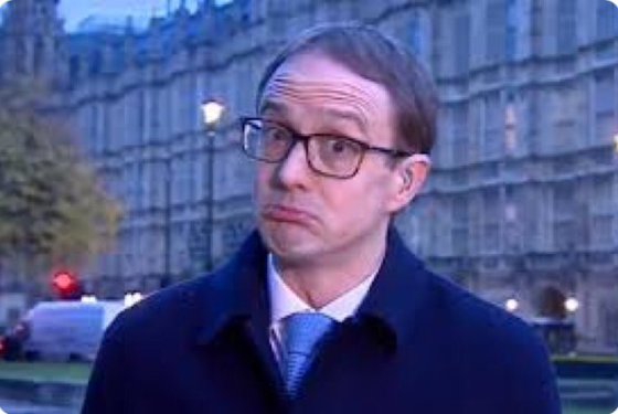 Man with glasses, dark coat and light blue tie standing outdoors in front of a large gothic stone building.