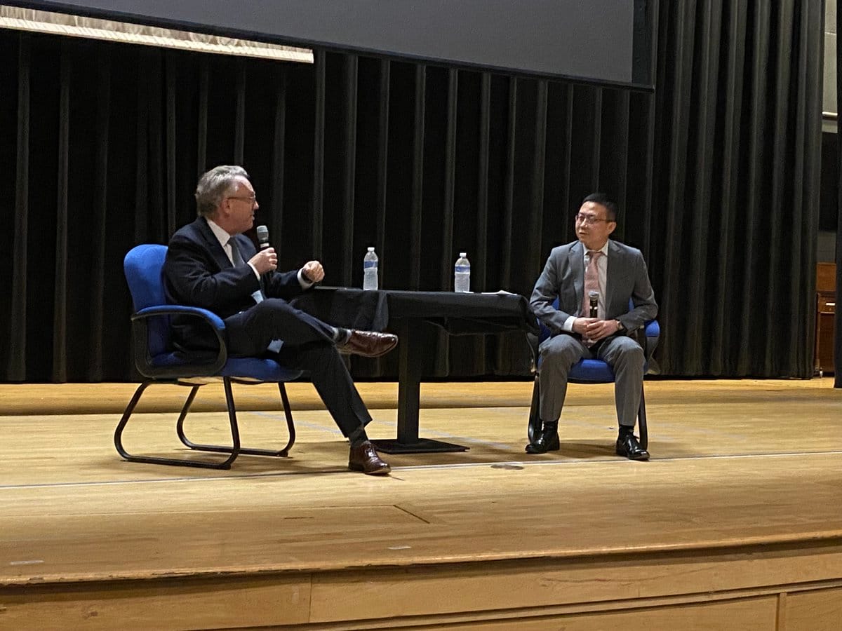 Two men in suits sit on a wooden stage with a small table and water bottles; the man on the left holds a microphone.