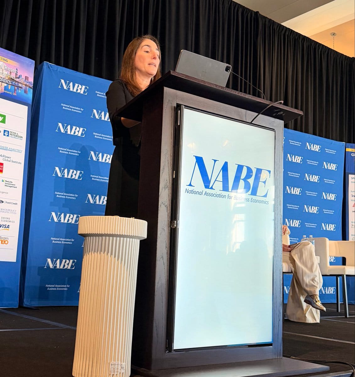 Woman speaking at a dark wood NABE podium with a laptop; blue NABE banners and a seated person in the background.