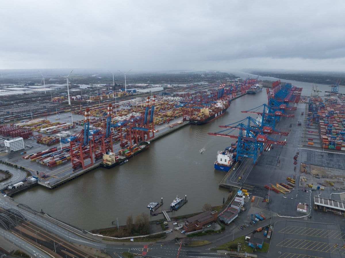 Aerial view of a container port with cargo ships, gantry cranes, stacked containers, and a canal under overcast skies.