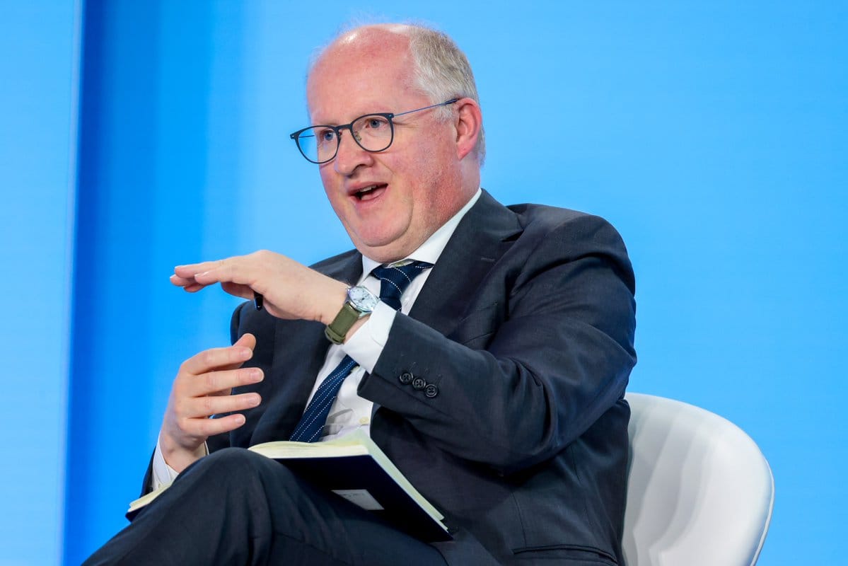 Balding man in glasses and dark suit sits on a white chair against a blue backdrop, gesturing while holding a notebook on his lap.