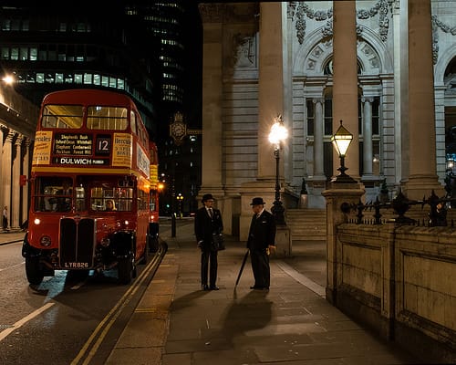 Night street scene with a red double-decker bus and two men in bowler hats on a London pavement beside a lit classical building.