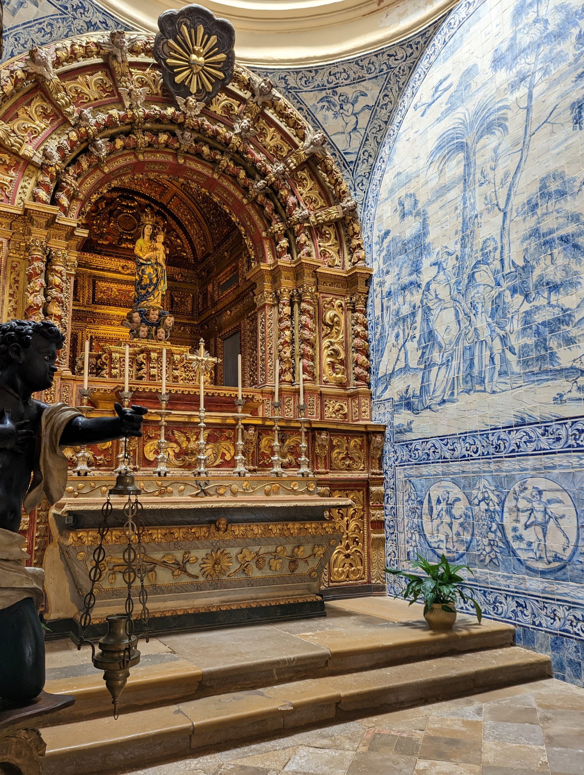 The scene inside of Faro Cathedral. A black angel is in the foreground, in front of a large, ornate golden altar with candles, a cross, and the Virgin Mary. The wall to the right is a religious scene created with Portugal blue and white tiles.