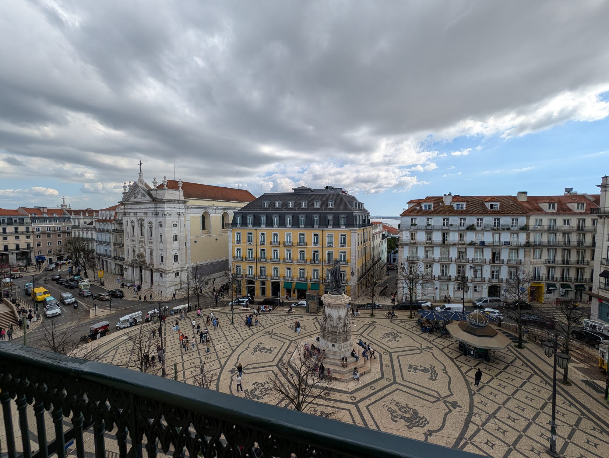 Looking from the balcony over a large plaza with a statue of the poet, Luis de Camoes. The plaza design includes important symbols and geometric tiling surrounding it. Surrounding the plaza are busy streets and historic buildings.