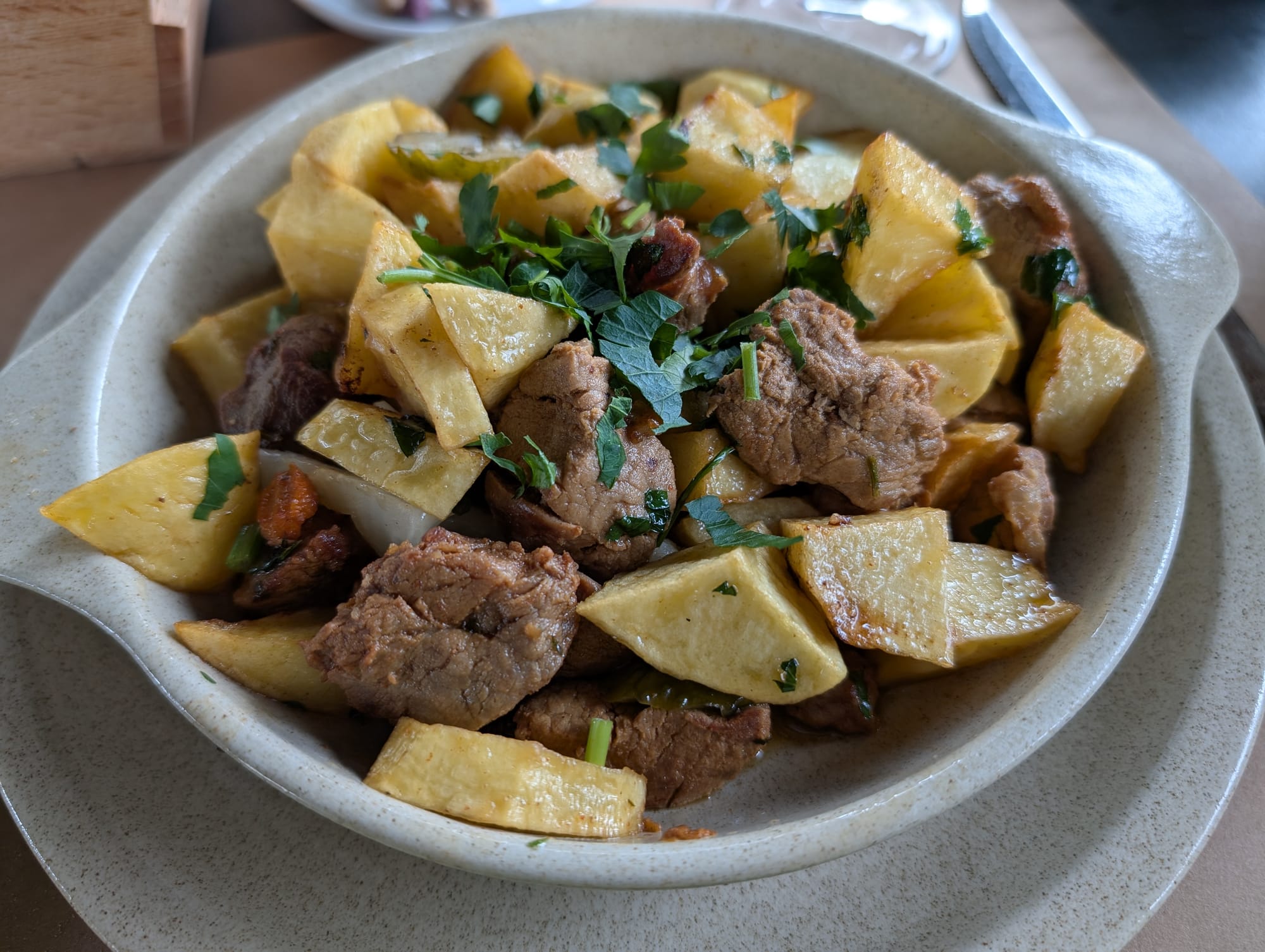 A ceramic bowl filled with pork, chunky potatoes, gherkins and herbs on top of a matching ceramic plate.