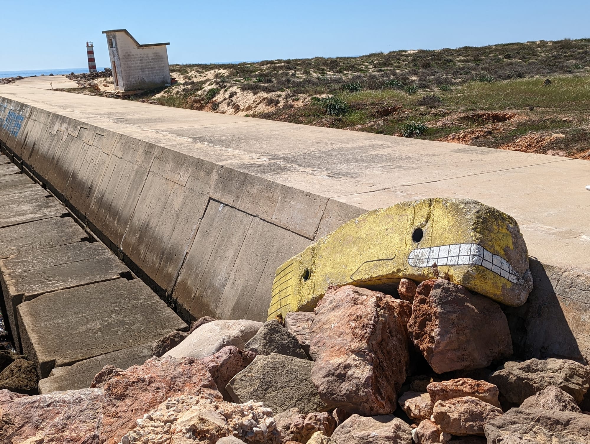 A yellow concrete whale atop a pile of rocks on a desert island, next to the shore. A lighthouse stands in the distance.