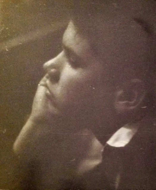Faint, faded, grainy vintage photo of a young boy in profile, with his chin resting on his hand.