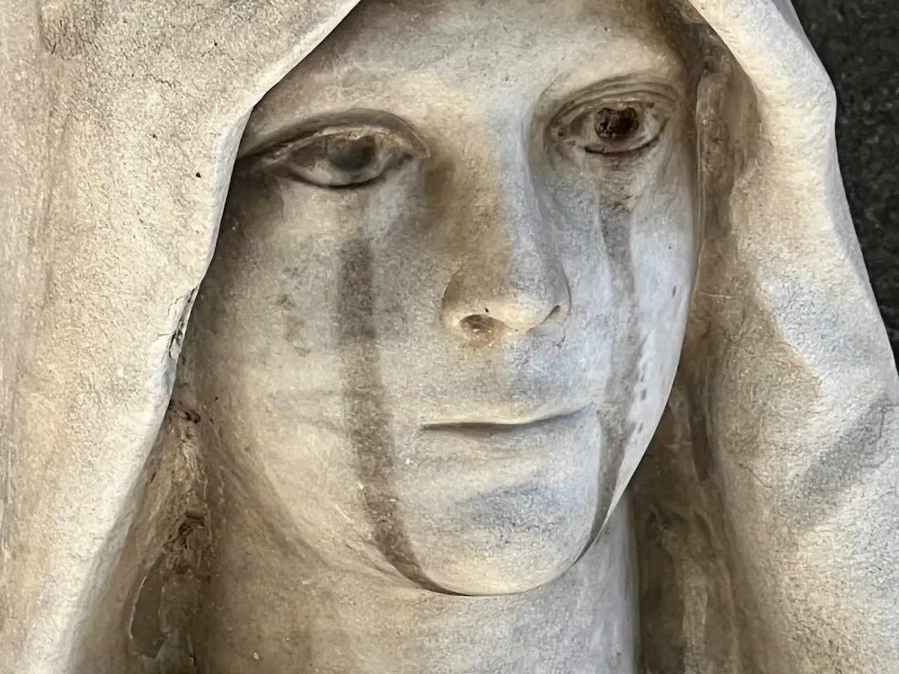Close up photo of a cemetery memorial sculpture of an androgynous face. There are stains running below the eyes that look like tear marks.