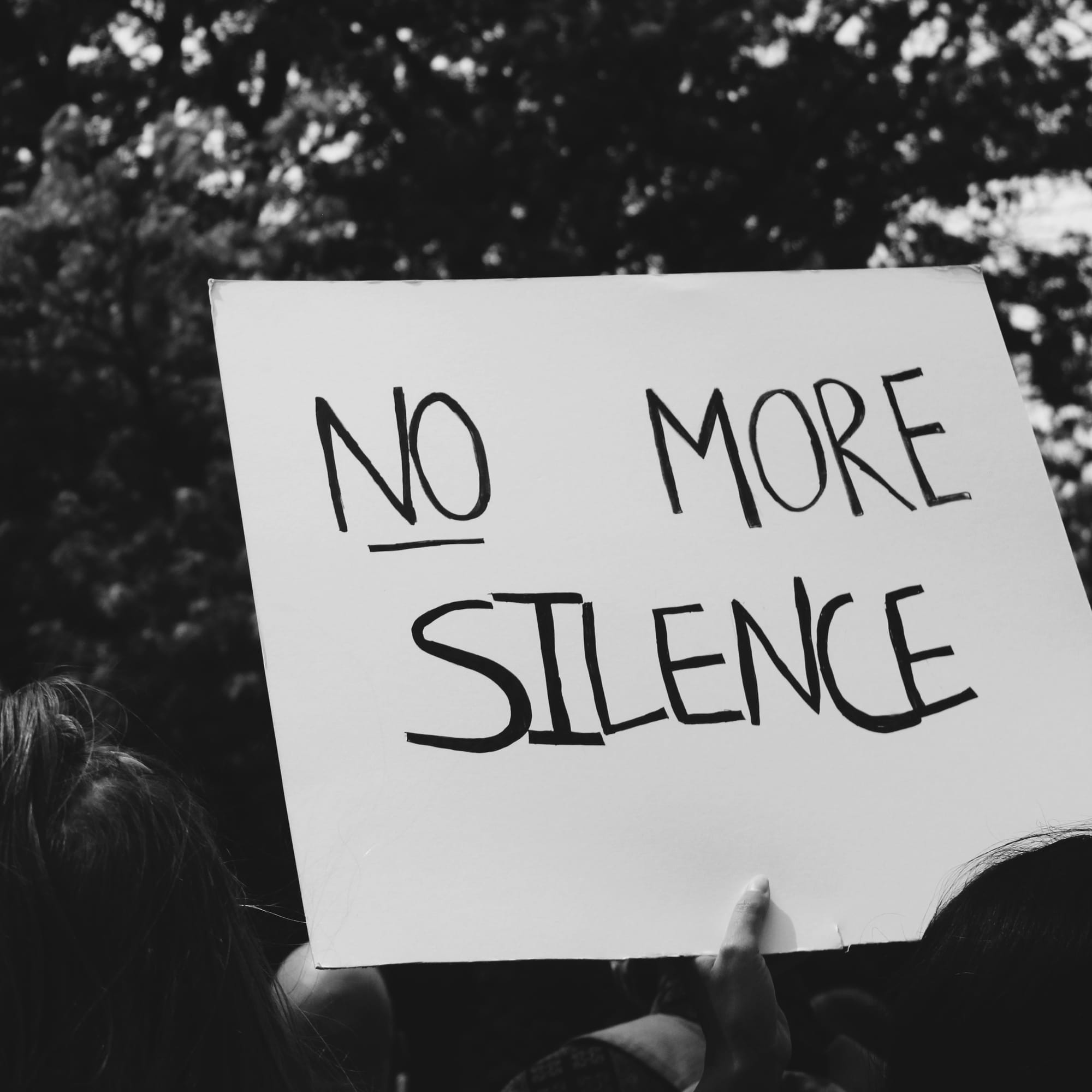 Photo of a hand holding a homemade poster with the handwritten words, "No More Silence."