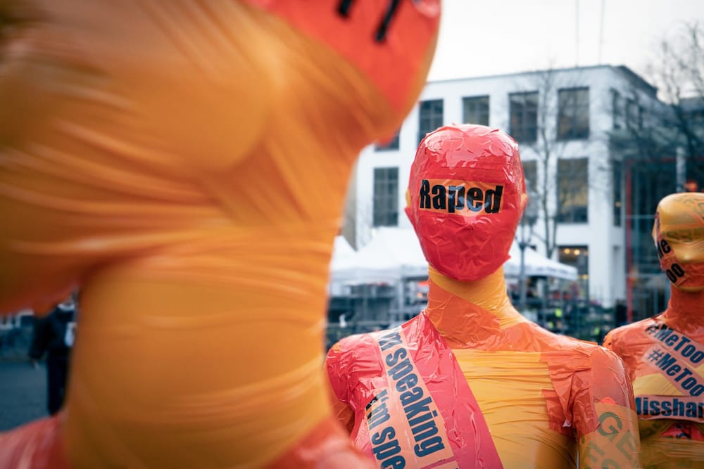Photo of three mannequins that have been wrapped completely in bright orange and red tape, with words written in black: "raped," and "me too."