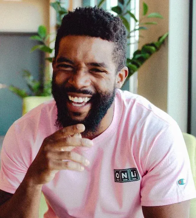 A black man with short hair and beard, and a big smile on his face. He is dressed in a pink t-shirt and looks like he's having a great laugh.