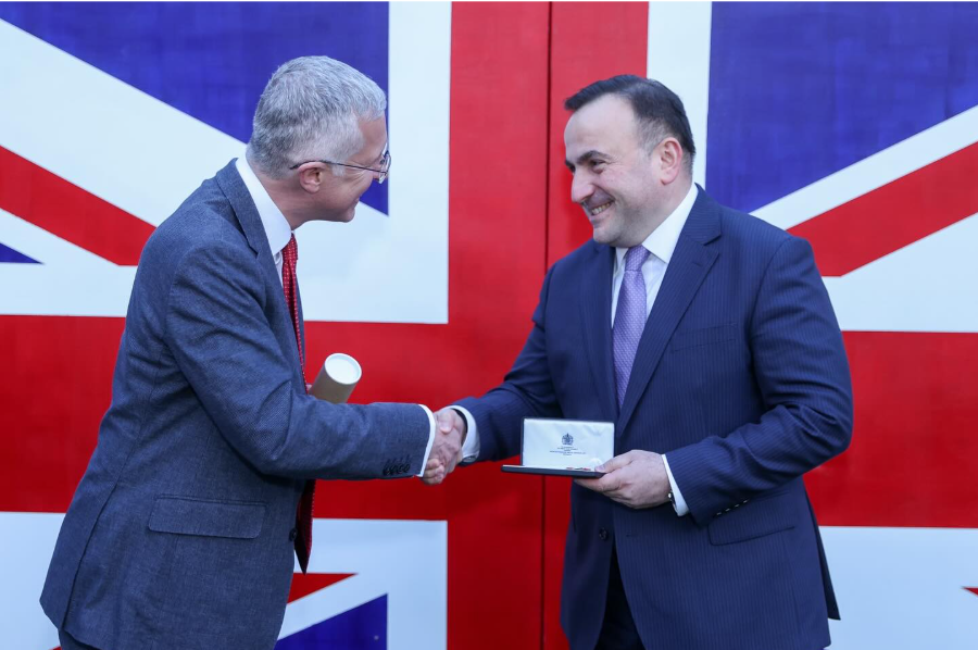 A grey-haired man in a grey suit shakes hands with a dark-haired man holding a medal box. The backdrop is a large Union Flag