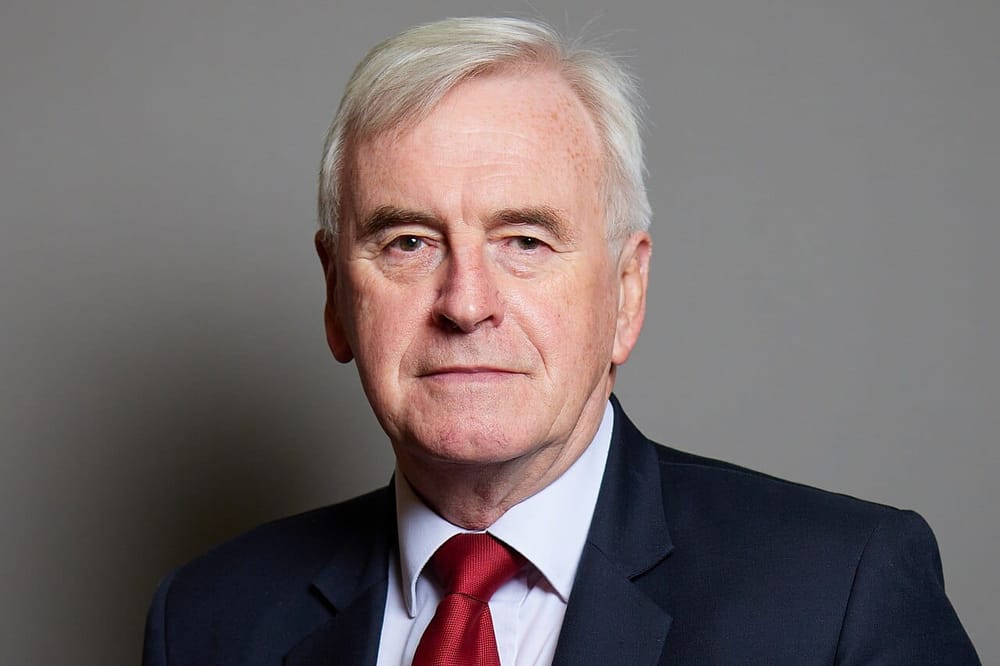 A man with grey hair, dark blue suit, light blue short, and red tie
