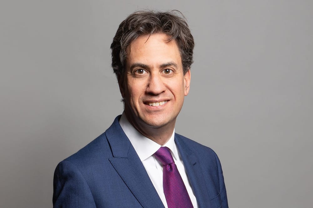 An official portrait of Ed Miliband, smiling, wearing a blue suit and purple tie